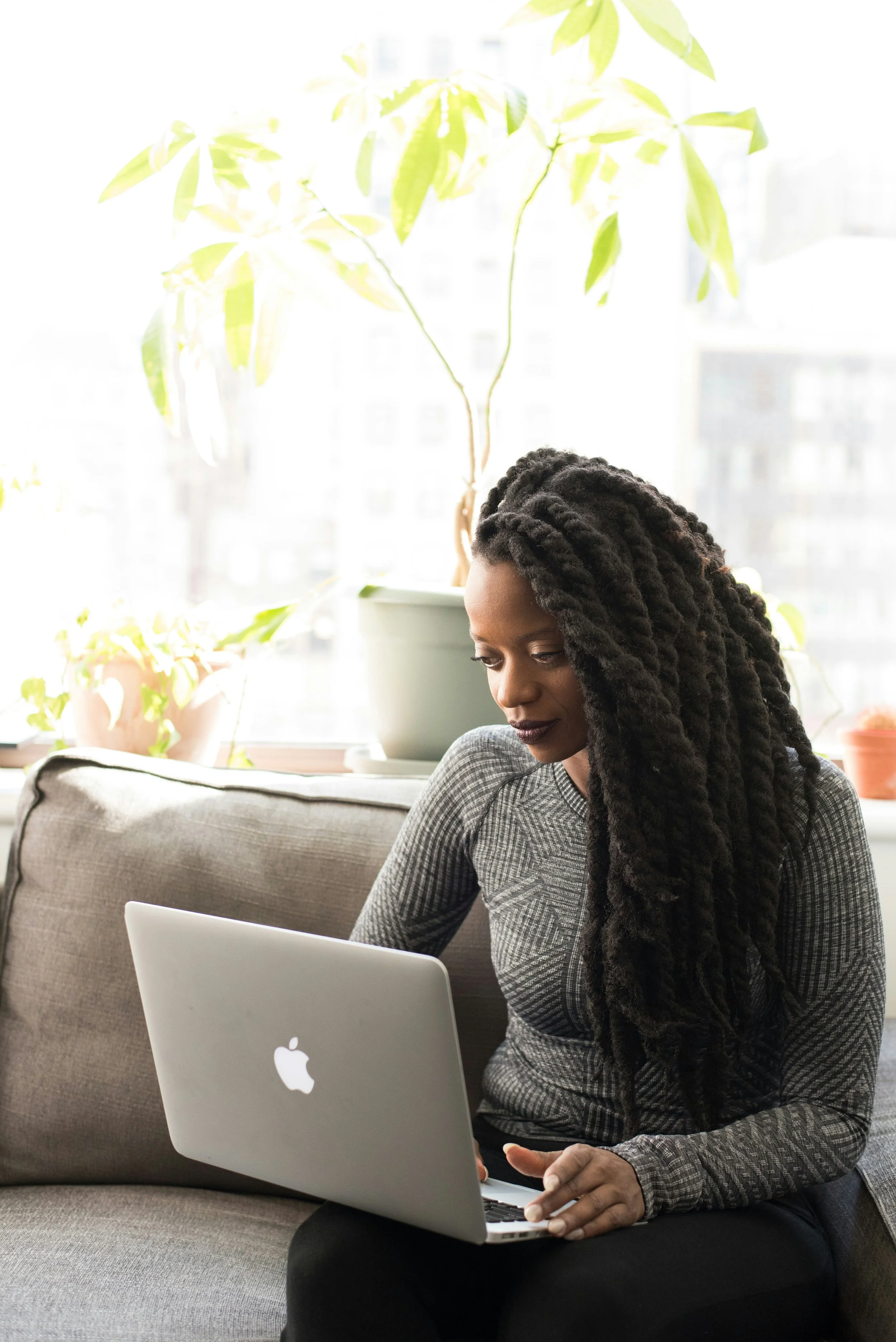 A woman on a sofa by a sunny window, browsing homes on her laptop