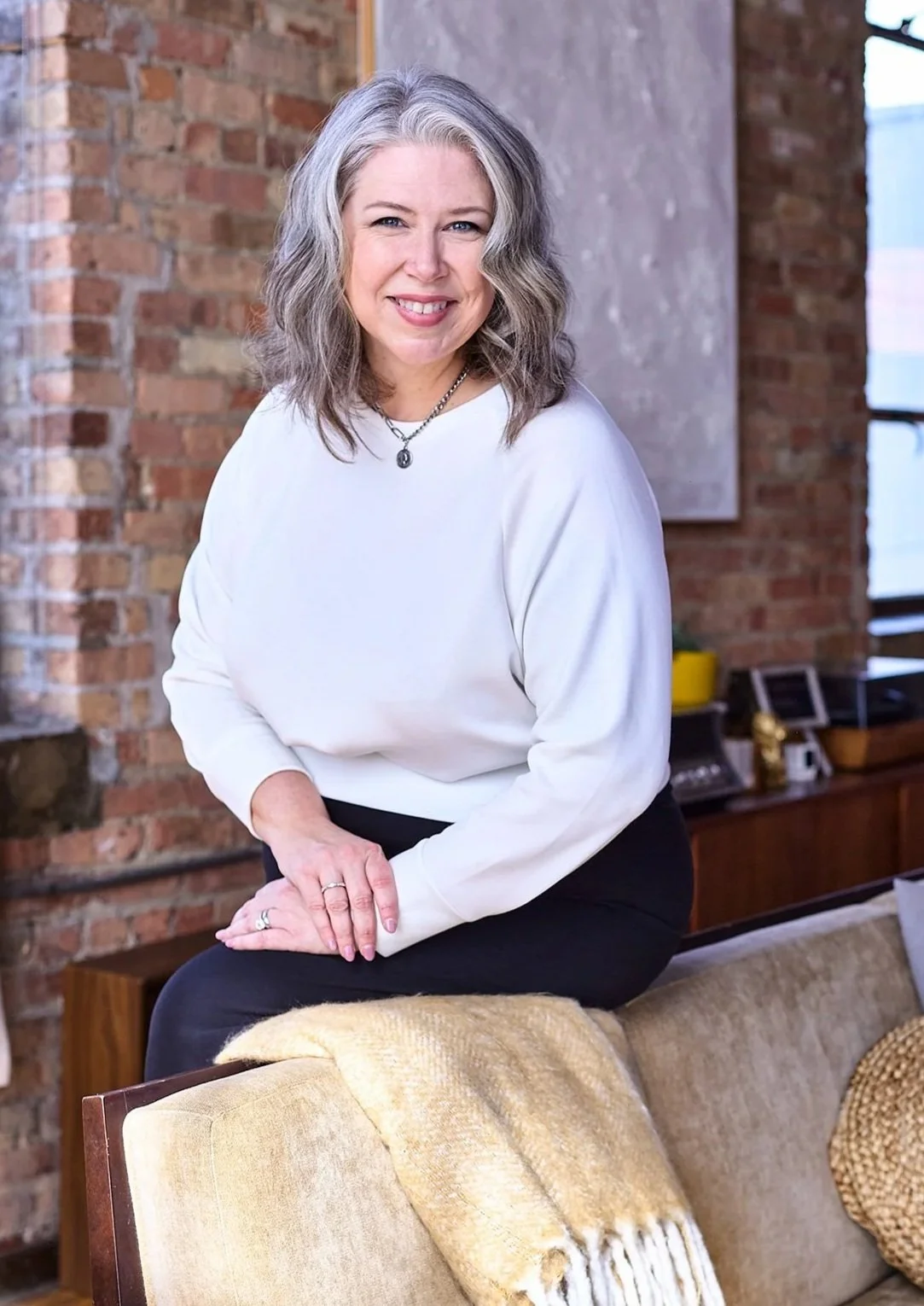 Deborah Libby, smiling, seated in a loft with exposed brick walls