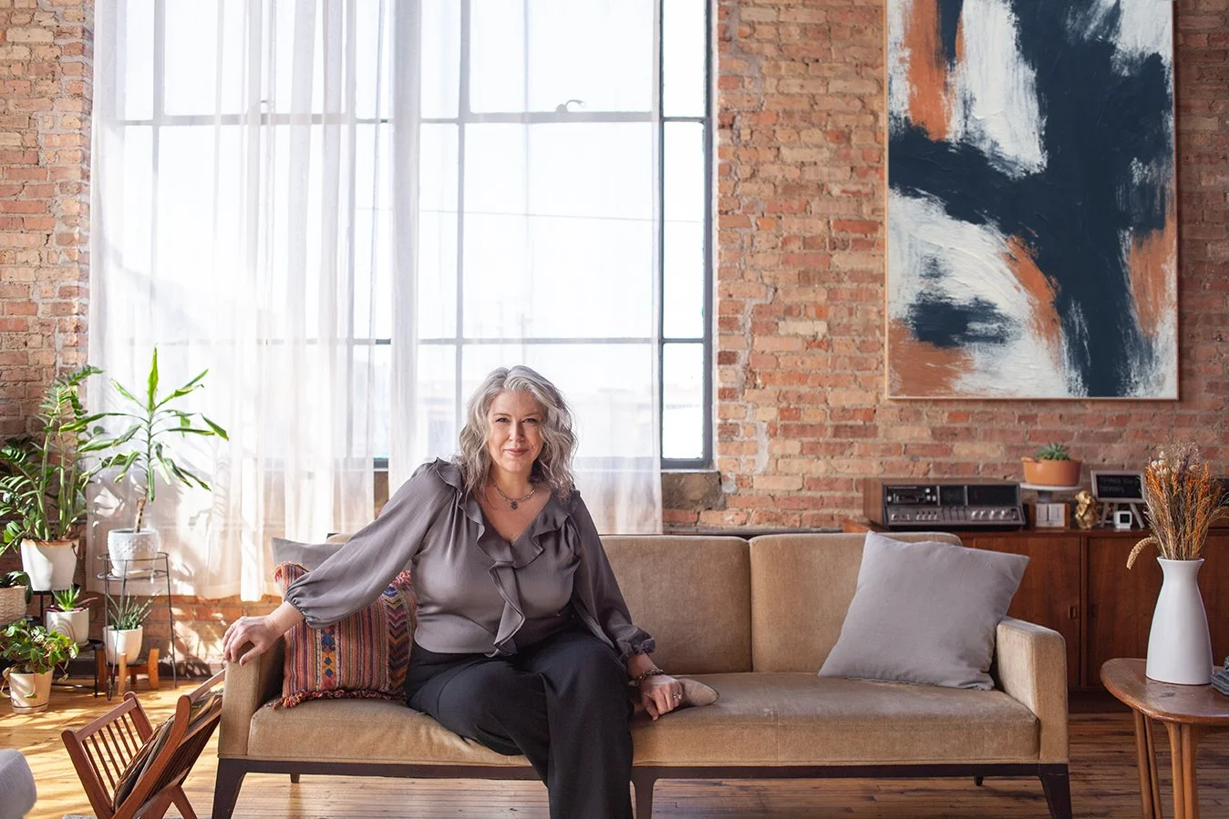 Deborah Libby seated on a sofa in a sunlit loft with exposed brick